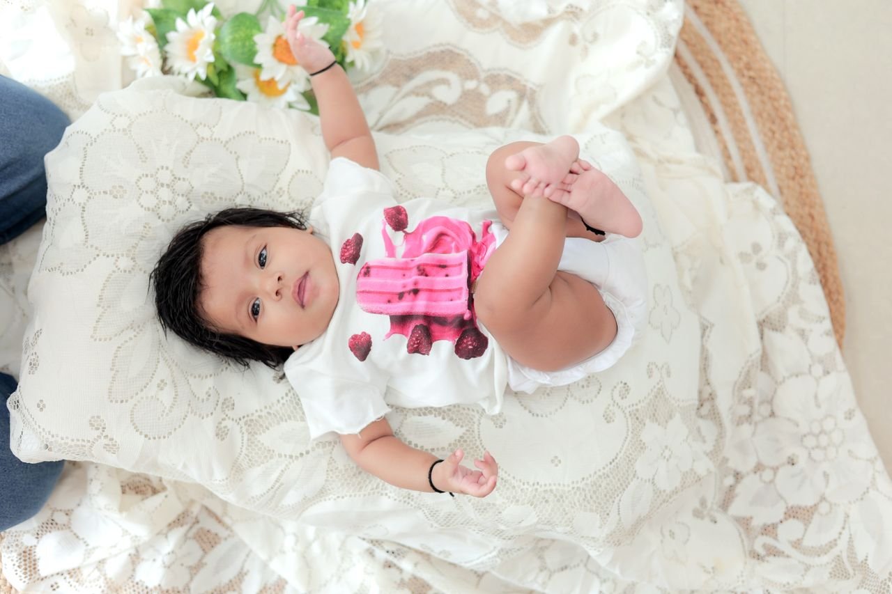 Cute baby lying comfortably on a floral lace-covered bed next to a bouquet of flowers, exuding innocence and warmth.