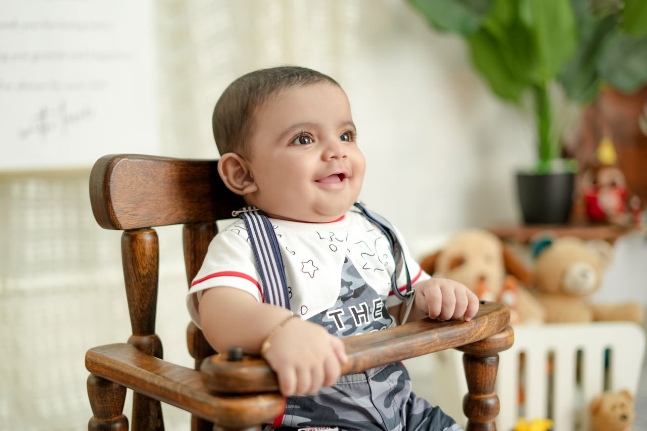 services-01 Smiling baby sits in a wooden high chair surrounded by toys in a bright indoor setting.